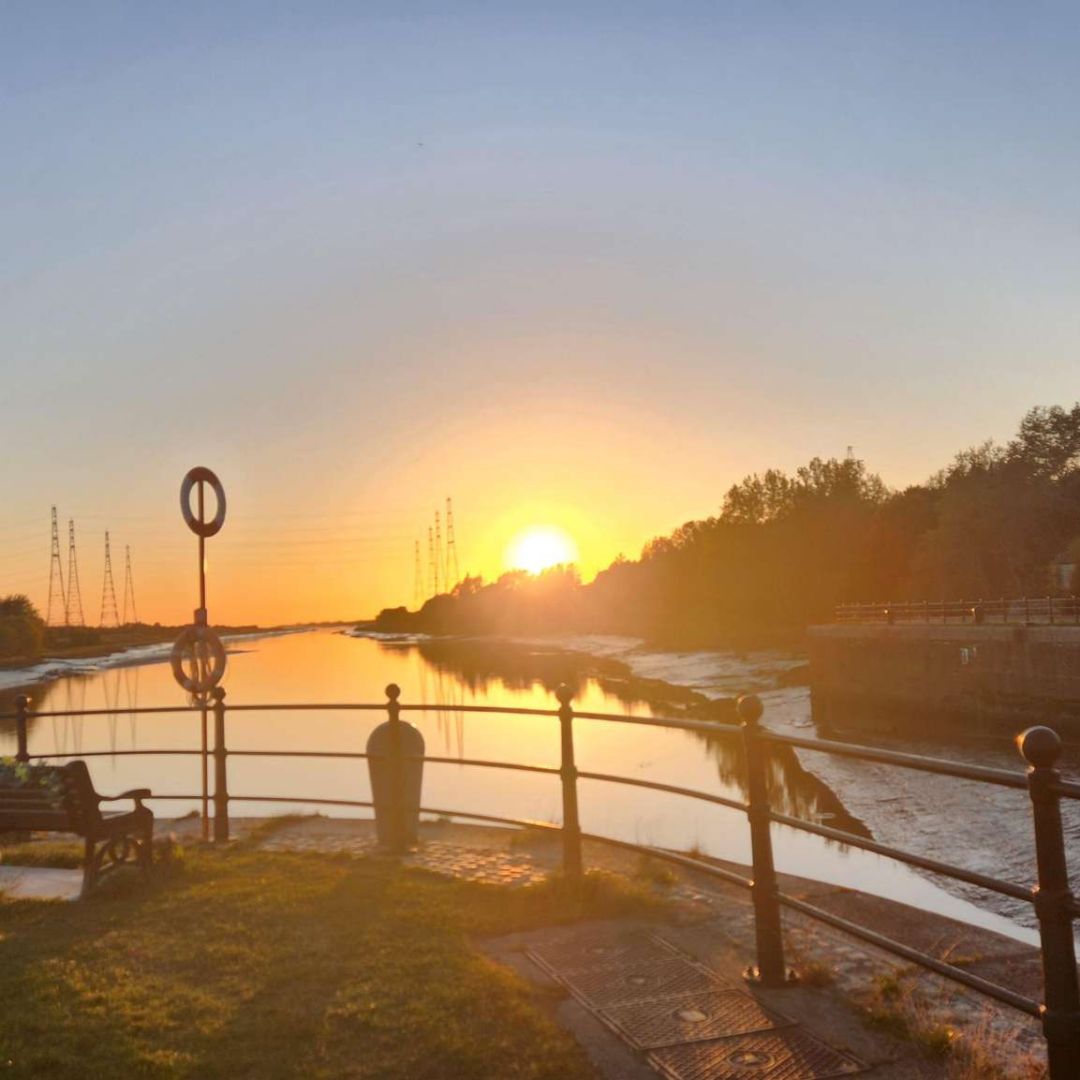 Sunset over Preston Docks Bullnose in Lancashire. The sun is low on the horizon, reflecting golden light across the calm water. Benches and railings line the edge of the dock, with trees and pylons silhouetted in the distance beneath a clear sky.
